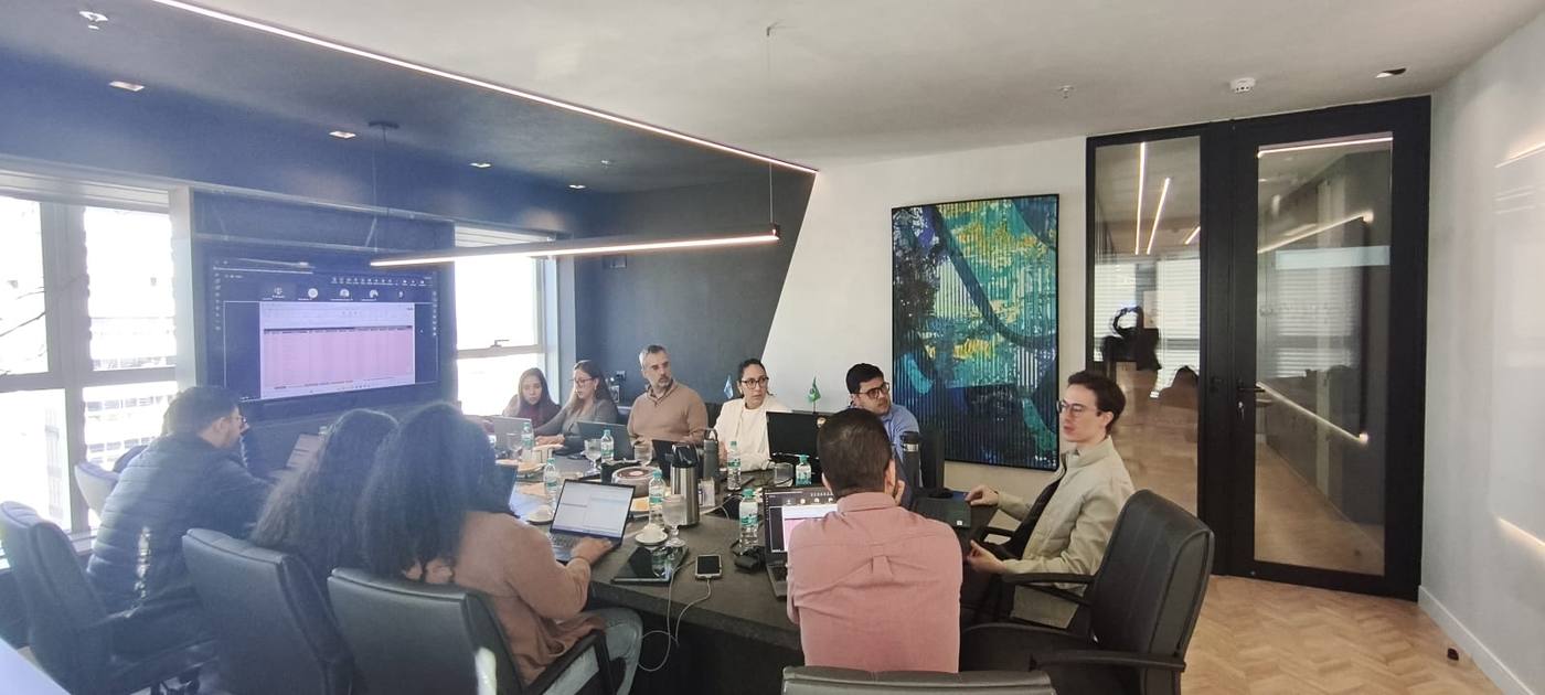 Sessão de trabalho da maestr·IA com o time de uma empresa cliente, em volta da mesa, com a planilha da operação projetada na tela